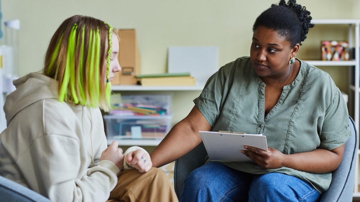A kind woman in her 40s is sitting with a teenaged girl who is not feeling well. The woman holds a clipboard in one hand and holds the girl's arm gently with the other.