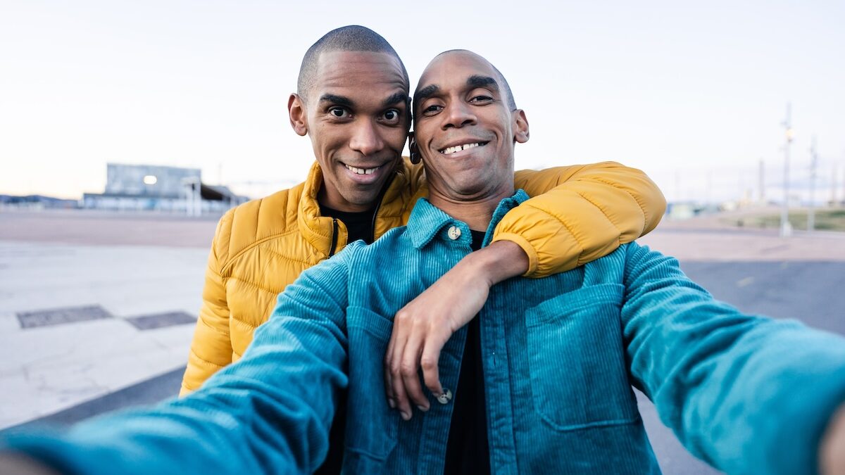 Smiling African American man in blue jacket taking selfie with his brother wearing yellow coat while standing together on city street during daytime